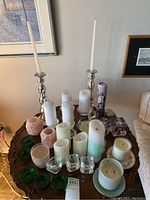 Wide view of various candles and candle holders arranged on a wooden table with art on the wall in background.