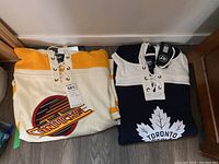 Folded Vancouver Canucks and Toronto Maple Leafs hoodie jerseys with visible tags laid on floor.