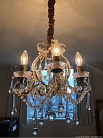 Front angle view of illuminated chandelier showing three candle-style bulbs, glass beads, teardrop pendants, and brown fabric covered hanging chain.