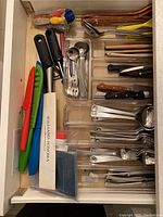 Overview of kitchen drawer contents showing knives with colored sheaths, flatware, peelers, and utensils organized in clear trays.