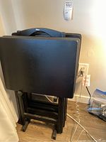 Side view of four black folding tray tables stacked leaning against the wall beside the wooden stand.