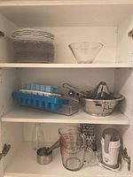 Shelving with 12 clear glass luncheon plates (3 made in Portugal) stacked on top shelf with a small glass bowl. Middle shelf has two blue ice cube trays, a metal handheld grater, metal strainer or sifter, and citrus juicer.