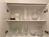 Overview of cabinet shelf showing 3 glass jugs (plain tall, embossed floral pattern, clear with lid), ceramic jug, gravy boat, small glasses and bowls