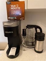 Keurig coffee maker with box of Van Houtte pods on top, thermos, Bodum frother, and Hamilton Beach kettle visible on countertop.
