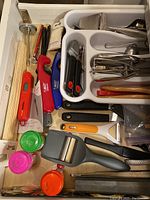 Photo showing an assortment of kitchen utensils laid out in a drawer organizer. Includes peelers, can opener, handheld graters, plastic measuring spoons, skewers, and various small tools.