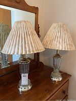 Two crystal base lamps with pleated beige fabric shades, set on a wooden surface with a mirror behind.