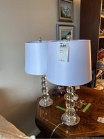 Pair of table lamps with glass raindrop bases and white drum shades sitting on a wooden table with an assortment of small items and paintings on the wall behind.