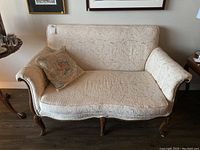 Front view of cream-colored upholstered loveseat with floral embossed fabric and decorative pillow on left side.