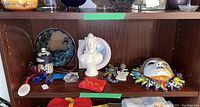 Shelf display of decorative bust, ceramic sun wall art, two plastic birds, decorative plate, small lizard figurine, red dish, and other small items.