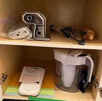 Photo of OXO mandoline slicer, Brita water filter jug, plastic cutting boards, and pasta drying stand parts in cupboard.