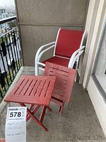 Photo shows set of 2 white metal framed outdoor chairs with red mesh seats and backs along with 2 red wooden slatted folding tables on balcony.