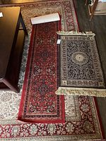 Three rugs displayed, one small rug with black and cream medallion and fringe, two runners with red and dark patterns, laid on larger cream and red patterned carpet.