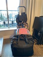 Rolser shopping trolley with geometric patterned bag, pink and turquoise travel bag, and large black travel bag positioned on the floor near a window and chair.