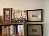Full view of three framed prints on wall and shelf, showing one tall ornate framed print, one square metal London/Paris print, and one small framed print.