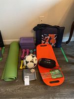 Photo of the green yoga mat, purple yoga blocks, various dumbbells in pink, yellow, and blue, white and black soccer ball, orange balance board, black football, fitness journal and navy blue bag