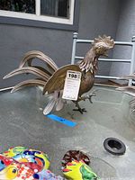 Standing brass chicken sculpture with open beak and raised tail feathers on glass table