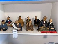 Full view of nine Hakata ceramic figurines arranged on a white shelf showing variety of traditional Japanese characters.