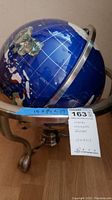Full view of the metal inlayed globe showing blue oceans and metal inlay continents, mounted on an ornate metal table stand with three legs.