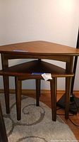 Two wooden nesting tables shown nested together, triangular tops with wood grain finish and straight legs