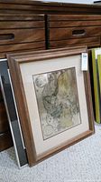 Side view of three framed pieces of wall art leaning against a wooden cabinet. Visible upper framed map with wood frame and matting.