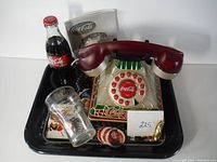 Photo showing Coca-Cola rotary dial telephone, branded glass, and small memorabilia items on black tray.