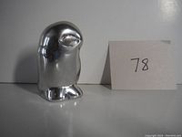 Front view of silver-tone metal owl sculpture with smooth surface and silver shine, alongside a paper card with number '78'.