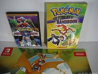 Photo showing Pokemon Destiny Deoxys DVD, Pokemon Ultimate Handbook book, and Nintendo Switch promotional sheet/map with Charizard and trainers.