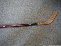 Full view of vintage wooden hockey stick featuring natural wood blade and Nike branding on black and red shaft, laid on carpeted floor.