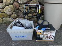 Photo showing white plastic bin holding various bicycle parts including Rock Shox Hydra Coil fork, handwritten parts, and package of tubes, alongside Pro Rac Bike Hoist bike rack box and Bell gel tech saddle on top