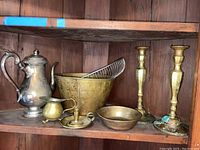 Photo showing silver plated coffee pot, brass bread basket with metal rack, brass candle holder with handle, two brass bowls, and brass candlesticks on wooden shelf.
