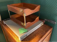 Photo of Ingento GT paper guillotine on wooden surface with two-tier wood and metal sorter trays labeled IN (top) and OUT (bottom). Portion of green wallpaper background visible.
