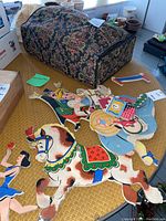 Photo showing multiple primitive puppets laid out on a woven surface including a large painted horse, a juggling woman, and other circus characters, along with a floral-patterned storage case.