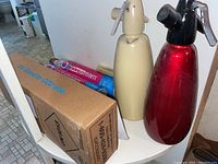 Two soda siphons, one beige and one red, with CO2 canisters and packaging on a small white surface