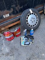 Photo showing four unused tires on Mercedes wheels, one used tire also mounted, two pairs of tail lights, car wash fluid bottle, wheel shine spray, and cleaning brushes arranged on ground.
