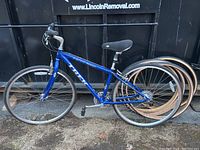 Side view of blue Trek 7300 Multitrack bicycle leaning against a black dumpster or container with wheels visible behind it.