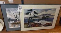 Two framed watercolor paintings leaning against a wall. One painting shows a tree with bare branches and snow, the other shows a lakeside scene with a tree and distant hills.