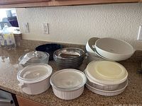 Photo showing metal mixing bowls stacked, glass casserole dishes with lids, plastic microwave bowls with lids, and ceramic casserole dishes on cabinet counter.