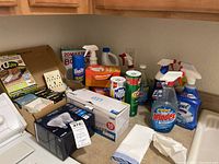 View of various used cleaning products on a kitchen counter including sprays, refill bottles, dryer sheets, and some packaging for bulbs.