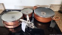 Six pieces of copper-colored cookware including frying pan, saucepans and a deep pot with glass lids. One lid is mismatched with a silver knob instead of copper.