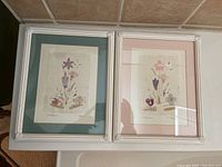 Two signed botanical prints shown in white frames with pink and green matting against a bathroom wall near window blinds.