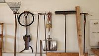 Photo showing an assortment of garden tools hanging on a wall including rakes, shovels, a push broom and a coiled hose