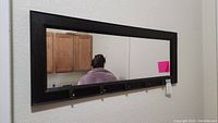 Full view of rectangular mirror reflecting a person with five coat hooks visible along the bottom edge of the dark frame.