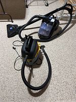 Two portable vacuums placed on carpeted floor. One is black with a clear container and taped wand attachment, the other is a gray and yellow cylindrical wet/dry vacuum.