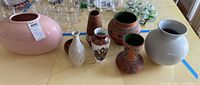 Wide angle image showing six pottery pieces including the large pink vase, smaller white vase with floral design, brown vase, and four Mexican pottery vases with decorative patterns, on wooden table surface alongside glassware.