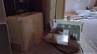 Singer portable sewing machine model 774 displayed with its matching cream color hard plastic carrying case. The sewing machine has dials for stitch types and speed adjustments and is on carpet flooring.