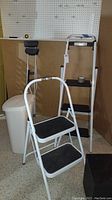 Wide view showing taller 54 inch stepladder, smaller two-step ladder, and plastic wastecan all positioned on carpeted floor against wall