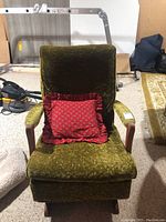 Front and angled views of the vintage crushed velvet rocking chair showing the olive green fabric, wooden armrests and base, and the red ruffled throw pillow.