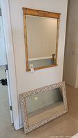 Full view of both wall mirrors, one gold-framed hanging on wall, other white framed resting on floor against wall.