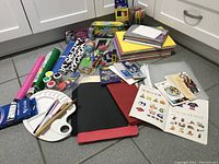 Overview of various art supplies including paper pads, paint, brushes, stickers, and other craft items arranged on floor