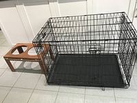 Photo showing black metal wire dog cage next to a rectangular wooden stand with two circular cutouts for bowls on tiled floor.